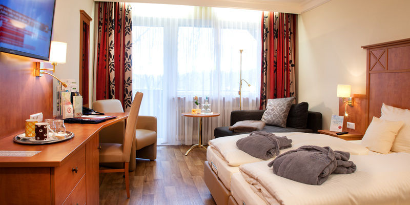 Modernes Komfortzimmer mit Doppelbett, Schreibtisch und Fensterfront vor der eine gemütliche Sitzgelegenheit steht.