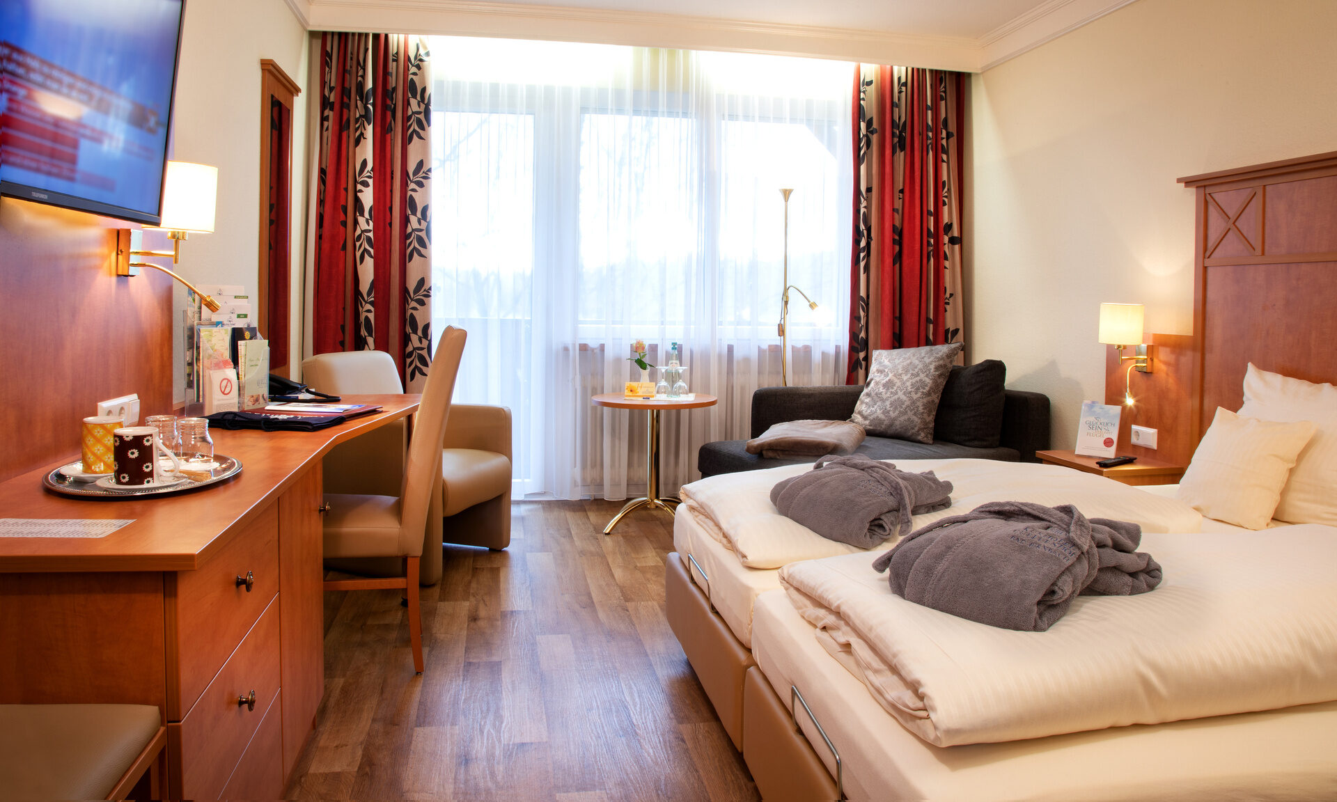 Modernes Komfortzimmer mit Doppelbett, Schreibtisch und Fensterfront vor der eine gemütliche Sitzgelegenheit steht.